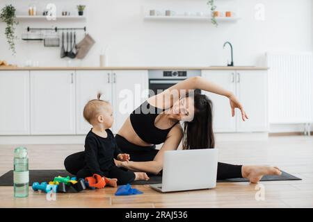 Wunderschöne junge Frau und lustiges kleines Mädchen, das auf Yoga-Matte neben Fitnessgeräten im Küchenhintergrund posiert. Fröhliche mutter, die sich seitlich beugt, während ein verspieltes Kind das gelbe Widerstandsband untersucht. Stockfoto