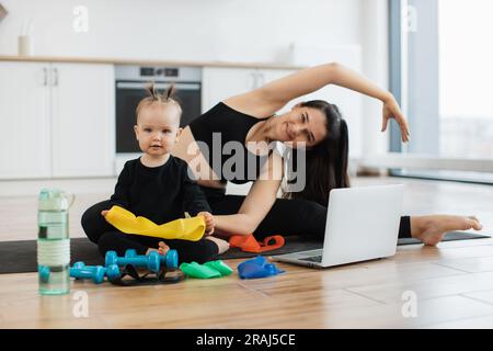 Wunderschöne junge Frau und lustiges kleines Mädchen, das auf Yoga-Matte neben Fitnessgeräten im Küchenhintergrund posiert. Fröhliche mutter, die sich seitlich beugt, während ein verspieltes Kind das gelbe Widerstandsband untersucht. Stockfoto