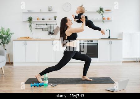 Sportliche Dame in Sportkleidung läuft durch einfache Dehnübungen, während sie das kleine Kind in Flugzeugposition hält. Sportliche weibliche Eltern verbessern die Koordination des Kindes beim täglichen Workout zu Hause. Stockfoto