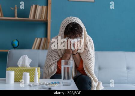 Ein kranker junger araber, der zu Hause auf dem Sofa sitzt und mit Papiertüchern niest. Männliche Nase bläst, Husten oder Niesen zu Hause, leiden an Grippe. Kalt Stockfoto