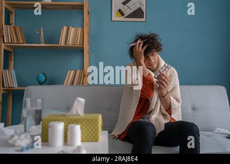 Arabischer junger Mann, der an Grippe und Erkältung leidet, hat Kopfschmerzen, während er zu Hause auf dem Sofa sitzt, das mit einer Decke bedeckt ist Stockfoto