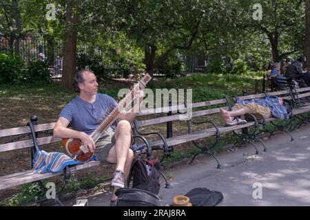 Eine New Yorker Straßenszene mit einem nicht-hinduistischen Sitarspieler in der Nähe eines schlafenden Schuhlosen. Im Washington Square Park in Greenwich Village, New York City. Stockfoto
