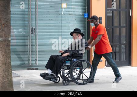 Ein ranghöchster chassidischer Jude im Rollstuhl, unterstützt von seinem Helfer. In einer ruhigen Straße in Brooklyn, New York. Stockfoto