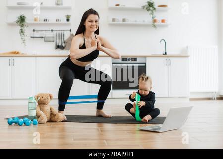 Eine charmante Frau in Aktivkleidung, die sich mit einem Widerstandsband duckt, während das Kind in der Wohnung ein grünes nimmt. Schlanke Mutter macht Übungen interessanter für neugierige Babys, während sie drinnen bleiben. Stockfoto