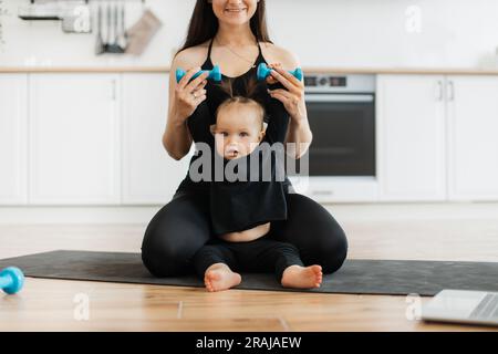 Freigestellte Ansicht einer Elternfrau, die die Hand des Babys hält und dabei hilft, die Kurzhanteln auf der Yoga-Matte im Innenbereich anzuheben. Süße kleine Tochter, die sich an einen aktiven Lebensstil gewöhnt hat, zusammen mit fürsorgender Mami zu Hause. Stockfoto
