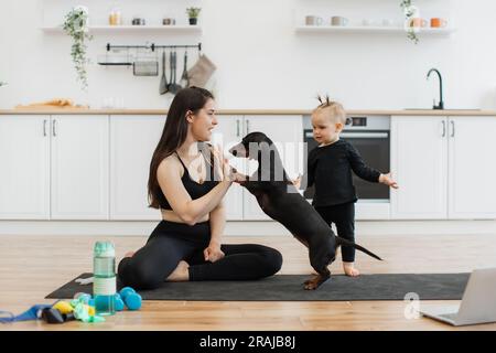 Hübsche Frau in Yoga-Kleidung gibt hgh fünf an kleinen Hund in der Nähe eines lustigen kleinen Mädchens in der Küche. Kaukasische Damen feiern effektives Training mit einem Tierfreund in der Wohnung. Stockfoto