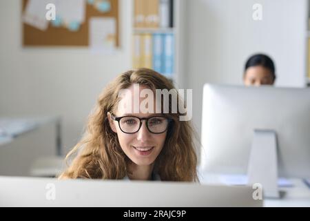 Junge Geschäftsfrau mit Brille, die am Tisch vor dem Monitor sitzt und am Computer arbeitet Stockfoto