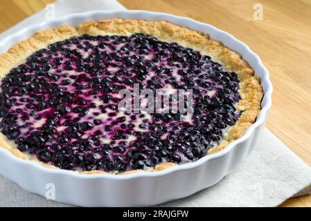 Hausgemachter Shortcrust-Gebäck-Kuchen mit Blaubeeren und saurer Sahne in einer weißen Keramik-Backform. Kuchen auf dem Holztisch und Leinentuch. Stockfoto