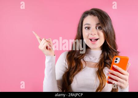 Mit einem Lächeln erregt eine reizende Frau Aufmerksamkeit, wenn sie das Kopierfeld zeigt und ein Mobiltelefon vor pinkfarbenem Hintergrund hält. Werbekonzept. Stockfoto