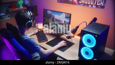 Ein junger Gamer kontrolliert den Tank, während er zu Hause am PC spielt. Computermonitor mit Live-Stream für Online-Videospiele oder Cyber-Sport-Turnier. Schreibtisch mit RGB-LED-Lichtband beleuchtet. Rückansicht. Stockfoto