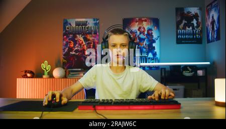 Junger Gamer mit Kopfhörern spricht mit Teamkollegen, spielt Online-Videospiele auf dem Computer im Neon-Raum für Cyber-Gaming. Aufgeregter Junge genießt Multiplayer-Cyber-Sport-Wettkämpfe. POV aus Sicht des PC-Bildschirms. Stockfoto