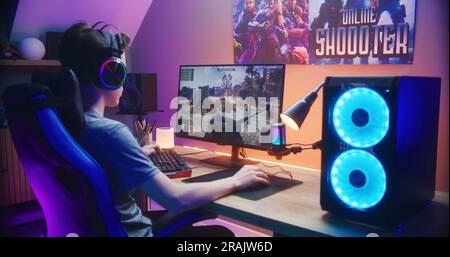 Ein junger Gamer kontrolliert den Tank, während er zu Hause am PC spielt. Computermonitor mit Live-Stream für Online-Videospiele oder Cyber-Sport-Turnier. Schreibtisch mit RGB-LED-Lichtband beleuchtet. Rückansicht. Stockfoto