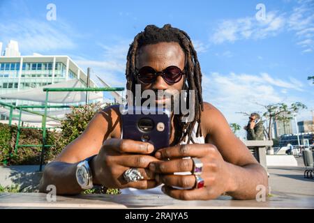 Vorderansicht eines jungen, gut aussehenden schwarzen Mannes mit Dreadlocks und Sonnenbrillen afrikanischer ethnischer Herkunft. Er sitzt draußen, telefoniert, surft im Internet. Stockfoto
