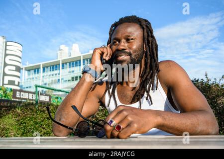 Junger afrikanischer Mann mit Dreadlocks und Bart, draußen in Buenos Aires, Argentinien, ruht sich aus und lächelt am Telefon, plaudert und chattiert Stockfoto