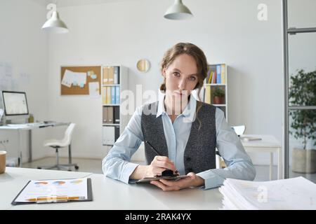 Porträt einer jungen Geschäftsfrau, die in die Kamera schaut, während sie sich Notizen in Dokumenten macht, die an ihrem Arbeitsplatz sitzen Stockfoto