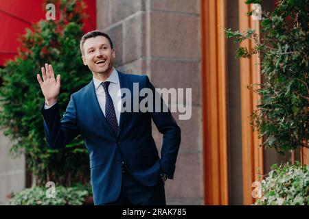 Fröhlicher Geschäftsmann, fröhlicher Gesichtsausdruck, formelle Kleidung, begrüßt Kollegen aus der Ferne. Im Freien in der Nähe des Restaurants. „Hey, ich bin hier!“ Stockfoto