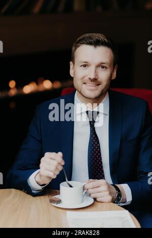 Zufriedener Mitarbeiter im Luxusanzug, trinkt Kaffee, lächelt, sitzt am Tisch, attraktives Aussehen, Wartet auf wichtige Gespräche mit dem Partner. Stockfoto