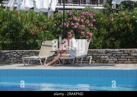 Touristenfrau in weißem Leinenmantel unter Sonnenschirm am Pool des Resorts, entspanntes Alleinurlaubskonzept Stockfoto