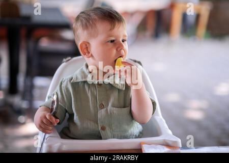 Süßer kleiner Junge, der Zitronenfrucht leckt, Ruhe im Hochstuhl und Löffel beim Abendessen Stockfoto