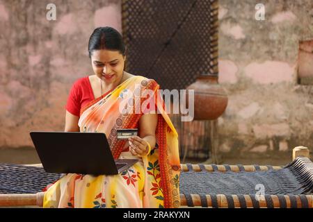 Glückliche Frau aus dem indischen Dorf in Saree, die den Laptop vor dem Haus benutzt und online mit Kreditkarte bezahlt hat. Stockfoto