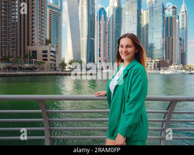 Junge Frau im grünen Anzug geht durch die Straßen von Dubai, Dubai Marina District. Reisekonzept der Vereinigten Arabischen Emirate. Die Vorstellung von erfolgreichem Auswanderer, Umzug in ein anderes Land, Arbeitsvisum Stockfoto