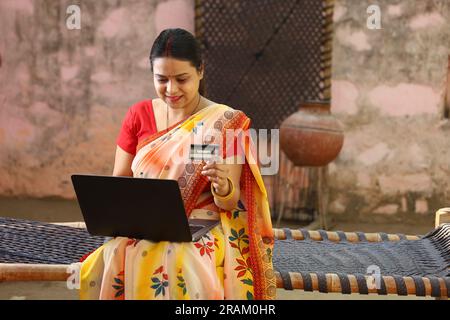 Glückliche Frau aus dem indischen Dorf in Saree, die den Laptop vor dem Haus benutzt und online mit Kreditkarte bezahlt hat. Stockfoto