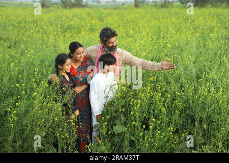 Glückliche indische Bauernfamilie, die tagsüber auf dem Senffeld steht und landwirtschaftliche Gewinne genießt. Sie freuen sich, von Senfpflanzen profitieren zu können. Stockfoto