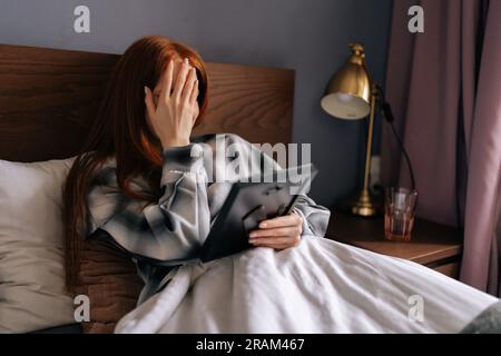 Seitenansicht einer trauernden jungen Frau, die auf dem Bett liegt und das Gesicht mit der Hand bedeckt und weint, während sie den Bilderrahmen hält und das Foto mit Liebe berührt. Stockfoto