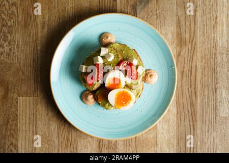 Blick von oben auf Avocado Toast mit Guacamole, weich gekochtem Ei, Frischkäse und Tomaten auf einem Teller. Stockfoto