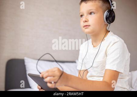 Lächelnder, glücklicher, weißer Junge im T-Shirt, der auf dem Bett sitzt und einen Tablet-Computer benutzt Stockfoto