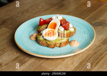 Avocado Toast mit Guacamole, weich gekochtem Ei, Frischkäse und Tomaten auf einem Teller. Stockfoto