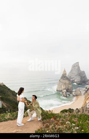 Sie hat oben "Ja" gesagt. Ein Paar, das auf einer felsigen Klippe am Meer steht, ein Mann, der der Frau einen Heiratsantrag macht Stockfoto
