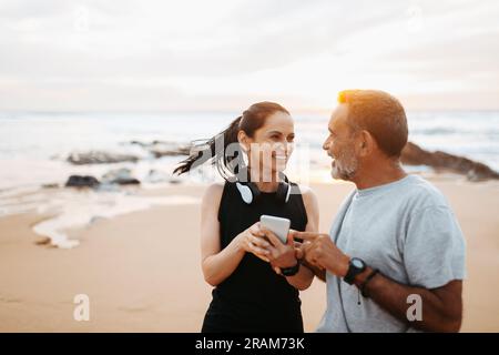 Die fröhliche ältere weiße Dame und der Mann in Sportbekleidung nutzen die Telefon-App, genießen Training und aktiven Lebensstil am Strand Stockfoto