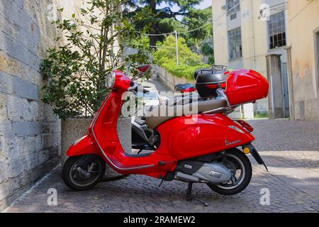 Ein roter Vespa-Roller parkt vor einer Kirche in der Stadt Belano in Norditalien. Sonniger Juni-Tag. Stockfoto