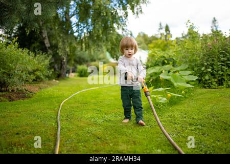 Süßer kleiner Junge, der am Sommertag im Garten Blumenbeete gießt. Kind, das Gartenschlauch zum Gießen von Gemüse verwendet. Kind hilft bei alltäglichen Aufgaben. Momm Stockfoto