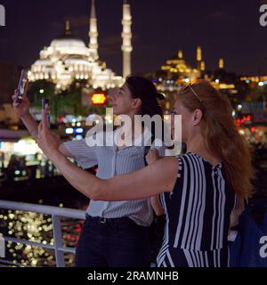 Zwei Frauen, Mutter und Tochter, nehmen nachts Selfies von einer Passagierfähre in Istanbul mit einer Moschee im Hintergrund. Stockfoto