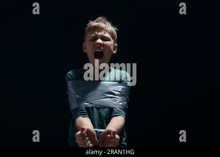 Ein vernarbter kleiner Junge, gefesselt und auf dunklem Hintergrund als Geisel genommen Stockfoto