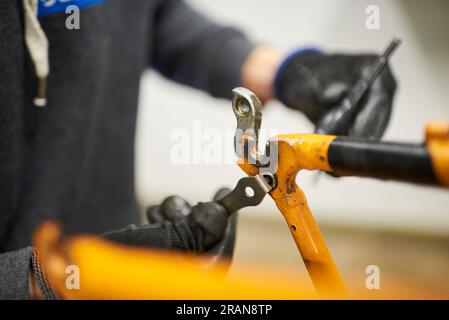 Reparatur eines Fahrrads: Hände einer nicht erkennbaren Person mit Handschuhen, die ein orangefarbenes Fahrrad in seiner Werkstatt demontiert. Selektive Fokuszusammensetzung. Stockfoto