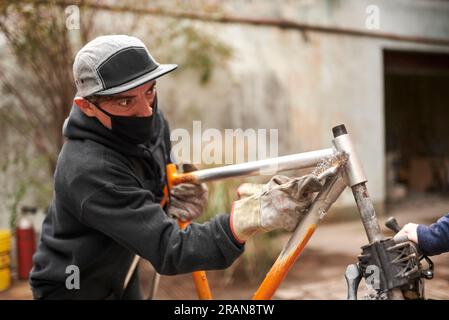 Junger spanischer Mann, der die Farbe eines orangefarbenen Fahrradrahmens entfernt, als Teil einer Fahrradrenovierung in seiner Werkstatt. Stockfoto