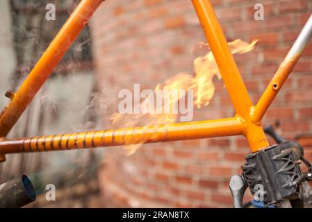 Eine nicht wiedererkennbare Person, die mit einem Lötbrenner die Farbe eines orangefarbenen Fahrradrahmens entfernt, der gerade in seiner Werkstatt renoviert wird. Stockfoto