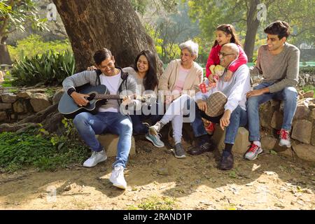 Happy Indian Family sitzt zusammen im üppigen grünen Park, spielt ein Lied mit Gitarre und genießt die Musik an einem Tag. Ruhig und friedlich Stockfoto