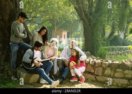 Happy Indian Family sitzt zusammen im üppigen grünen Park, spielt ein Lied mit Gitarre und genießt die Musik an einem Tag. Ruhig und friedlich Stockfoto