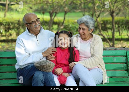 Großeltern sitzen auf einer Bank mit Enkelin im grünen Park, umgeben von ruhiger Atmosphäre und frischer Luft. Kein Verschmutzungstag. Stockfoto