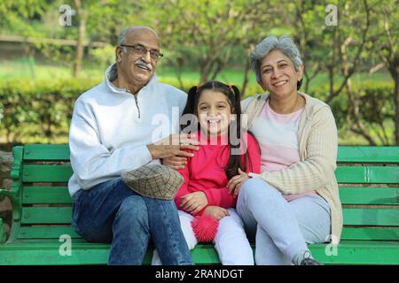 Großeltern sitzen auf einer Bank mit Enkelin im grünen Park, umgeben von ruhiger Atmosphäre und frischer Luft. Kein Verschmutzungstag. Stockfoto