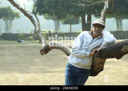 Gesunder, indischer, fröhlicher alter Mann im Park, der neben dem Baum steht und Spaß hat. Er trägt eine Golfmütze, die neben einem Ast steht. Stockfoto