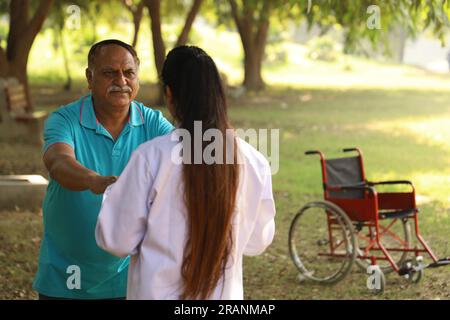 Weibliche indische Ärztin kümmert sich um Seniorpatienten im Krankenhausgarten. Der Patient ist gehunfähig Stockfoto