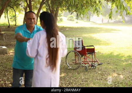 Weibliche indische Ärztin kümmert sich um Seniorpatienten im Krankenhausgarten. Der Patient ist gehunfähig Stockfoto