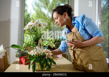 Wunderschöne, multiethnische Floristin, Blumendesignerin in beigefarbener Schürze mit Handy, die mit Blumenarrangements für arbeitet Stockfoto