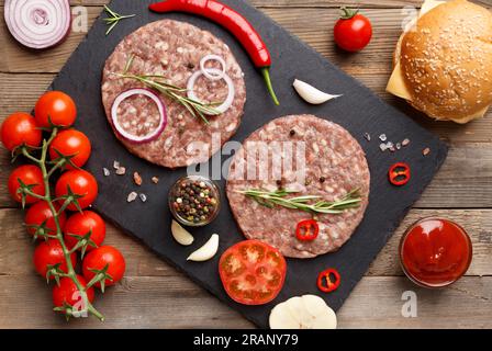 Burgerpasteten mit Rosmarin und Gewürzen auf schwarzem Schieferteller, frische Tomaten, Zwiebeln, Sesambrötchen, Käse. Stockfoto