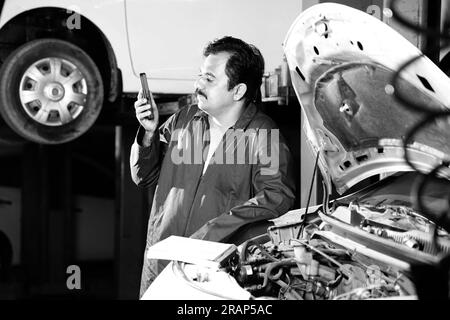 Ein denkender Automechaniker mit Schnurrbart, der seinen Tagesplaner hält, während er das Auto untersucht. Ein Autospezialist steht neben der offenen Motorhaube Stockfoto